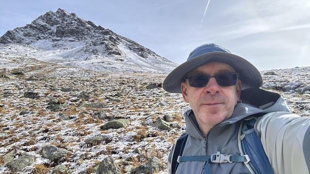 Selfie of me, a middle-aged white guy in sun hat, sunglasses, gray waterproof jacket and a backpack, in front of a rocky alpine slope with just a bit of snow on it.