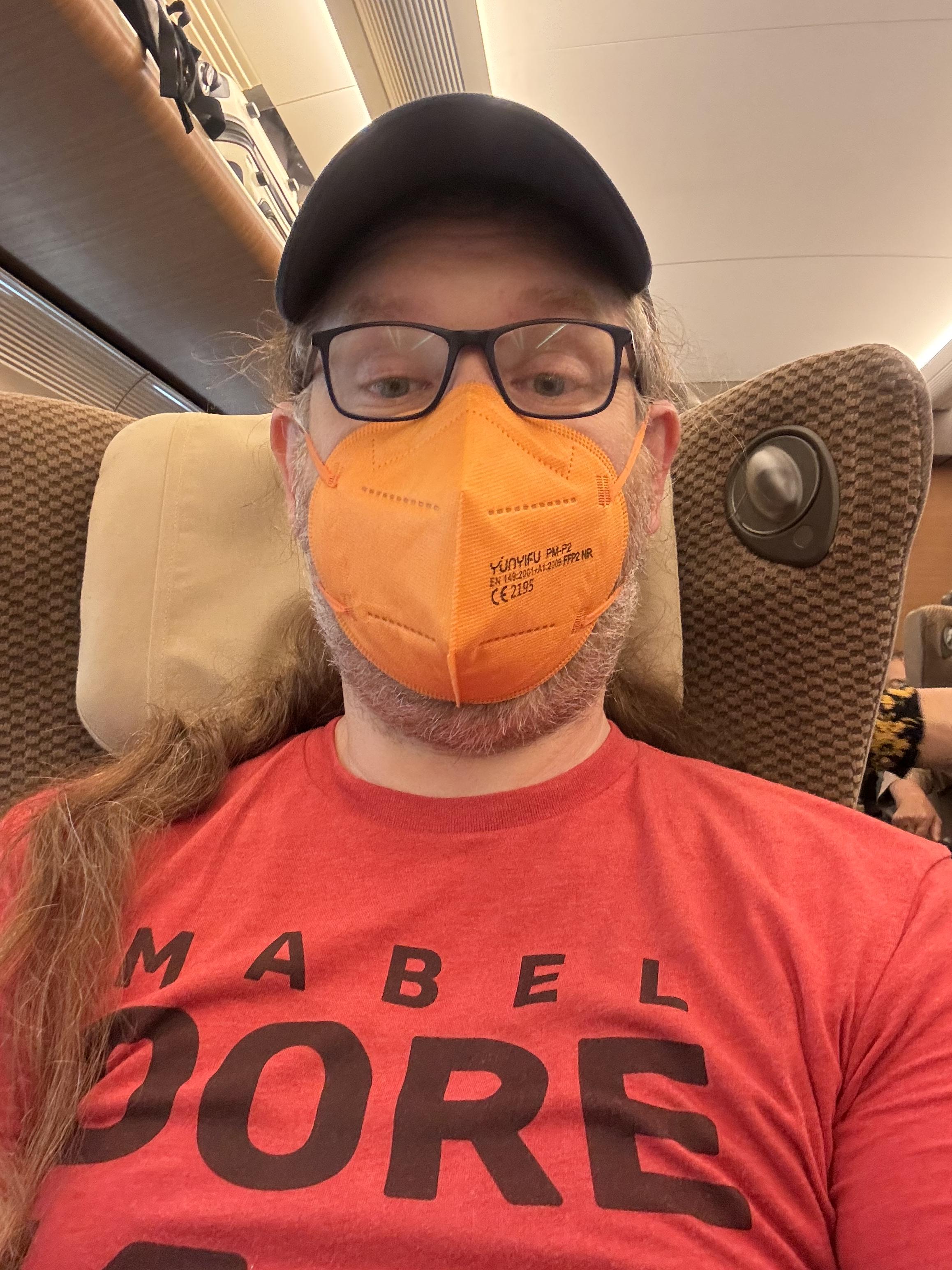 Selfie on train. Middle-aged man with a red tshirt, orange ffp2 mask, blue rimmed glasses and a blue basecap in a brown train seat, long hair casually flung over the left shoulder.