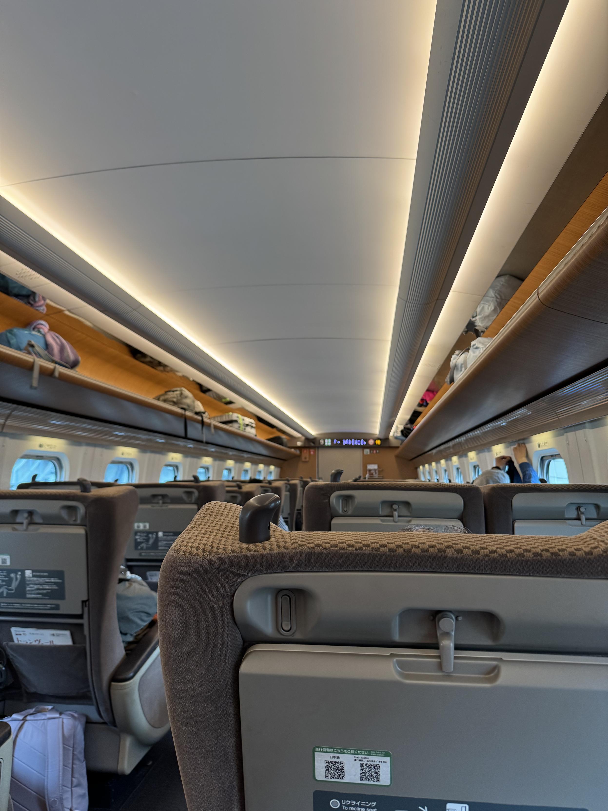 A Shinkansen train car with seats from behind, small windows, luggage racks and indirect lighting