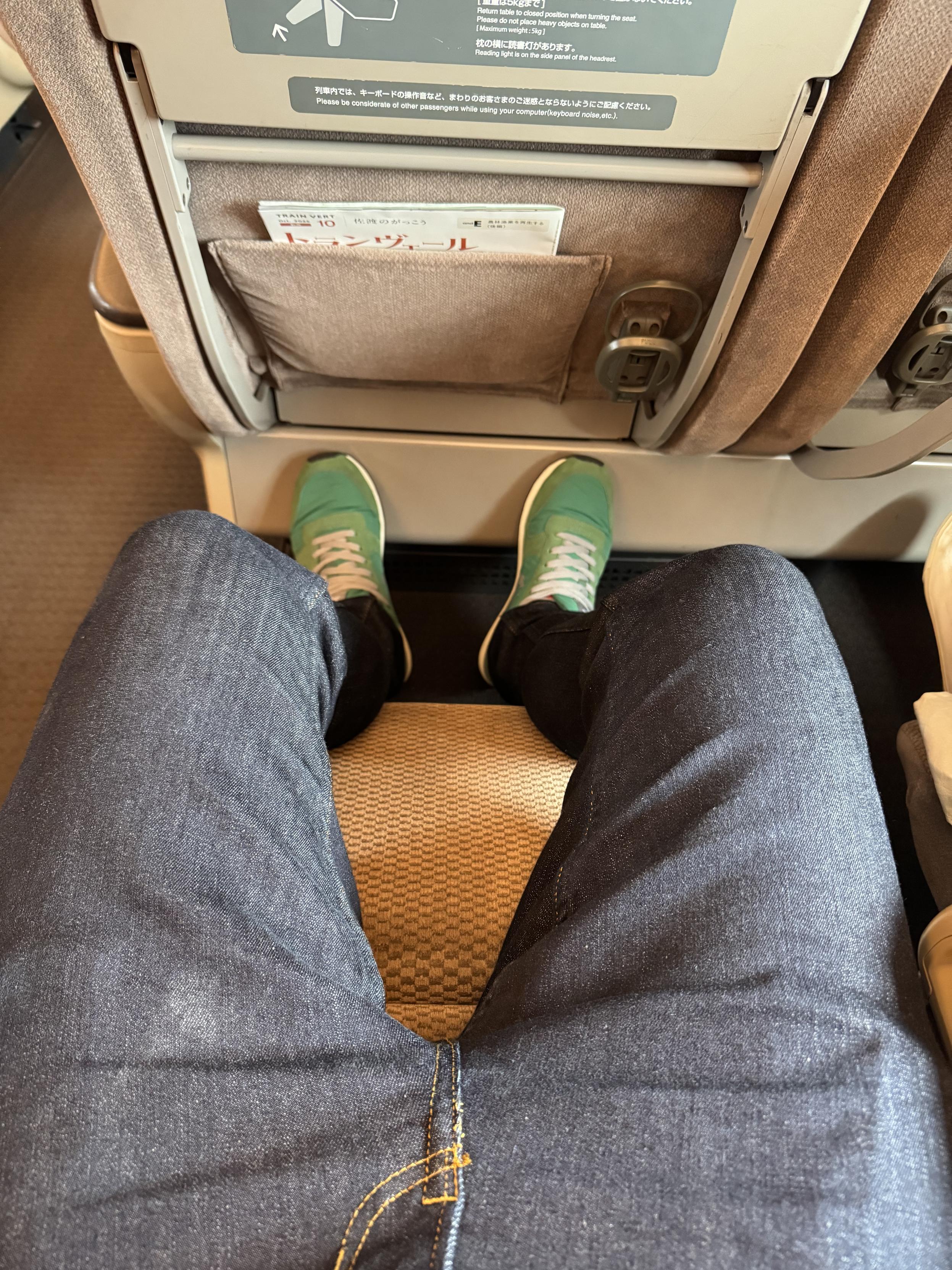 A tall person’s long legs in blue jeans and green trainers on a foot rest with with ample leg room