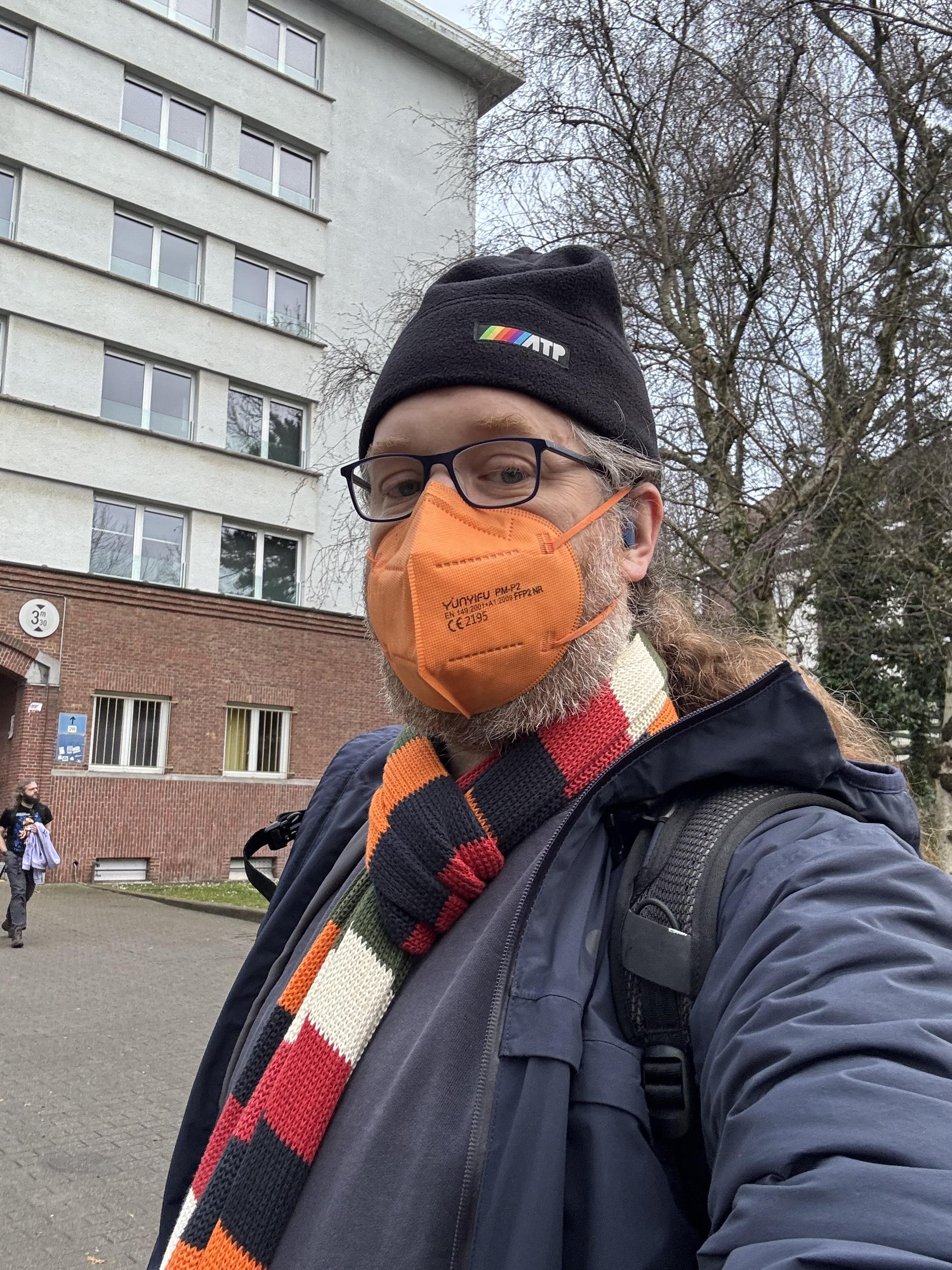 Selfie of a middle aged man standing in front of a university building. Wears a winter hat, jacket, striped shawl and bright orange ffp2 mask