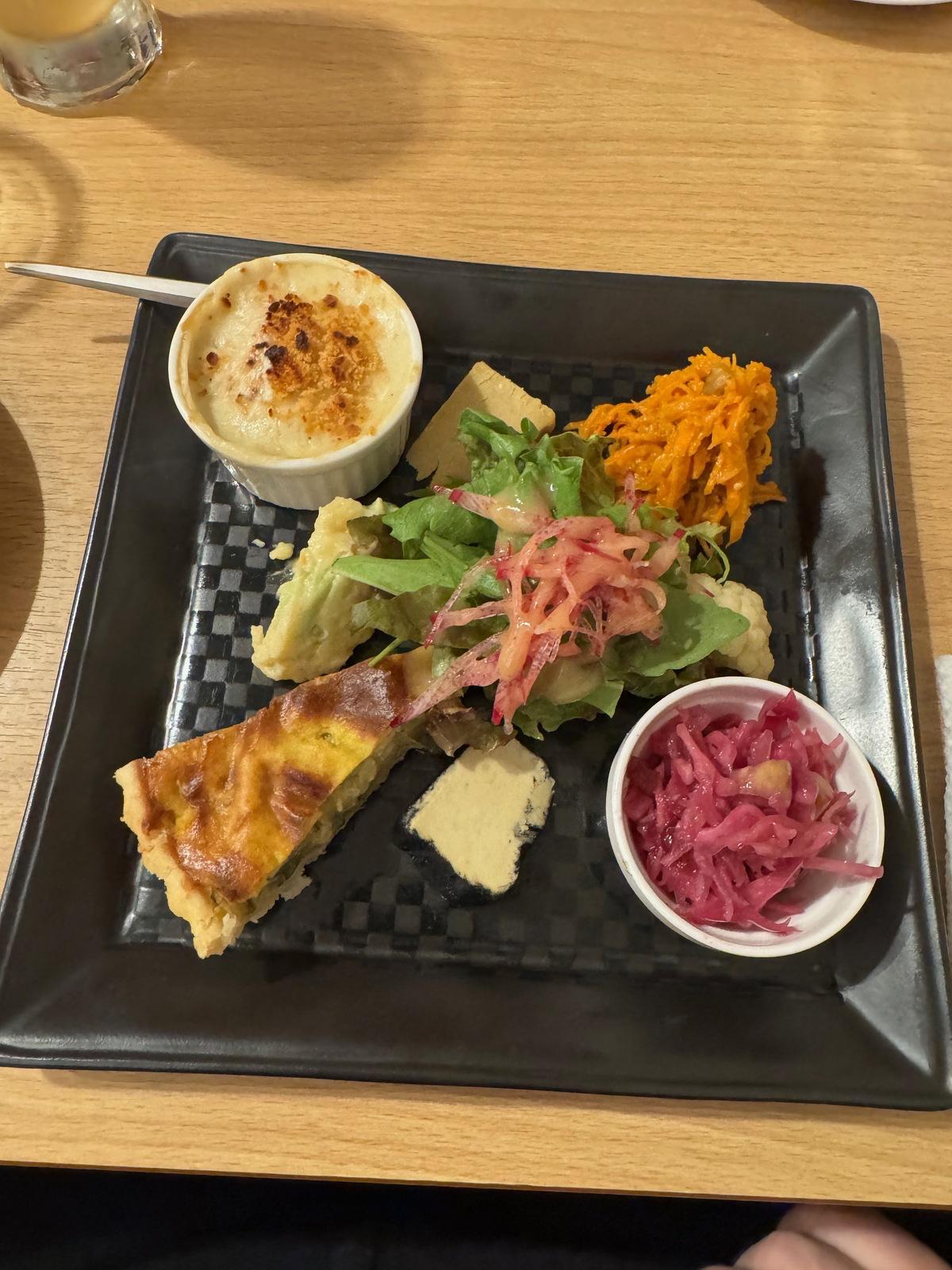 appetizer platter with quiche, carrot salad, vegan cheese slices, green lead salad, tiny mac and cheese bowl and pickeld cabbage salad